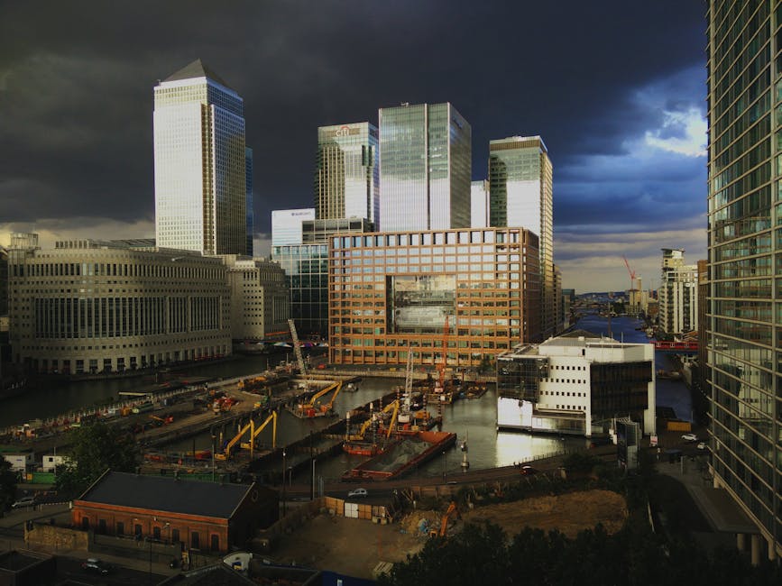 A wide-angle view of a modern cityscape featuring several tall office buildings with glass facades and a prominent orange-bricked structure in the center, situated next to a waterway with construction