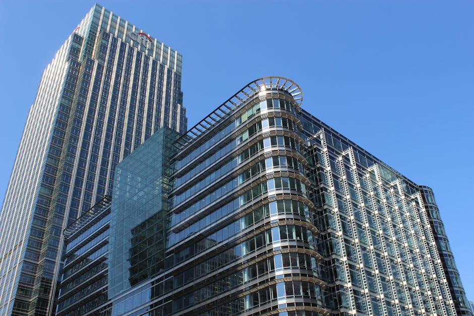 A wide-angle view of a modern cityscape featuring several tall office buildings with glass facades and a prominent orange-bricked structure in the center, situated next to a waterway with construction
